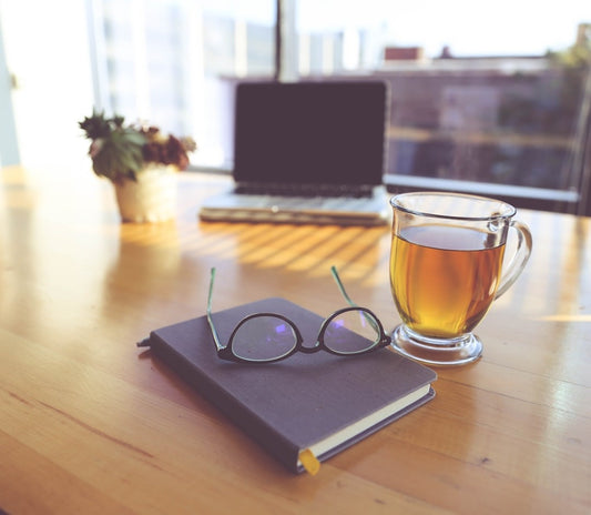 A photo of a desk facing a window with a journal, glasses, computer, bouquet of flowers and a cup of tea.