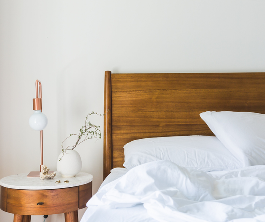 A bed with white sheets and a wooden headboard next to a wooden bedside table with a lamp and plant on the table.